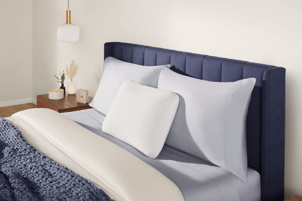 The TEMPUR-Symphony® Pillow placed on a stylishly dressed bed with a blue upholstered headboard, showcasing its premium design and adaptability for all sleep styles, available at Sleepology Mattress Shop.