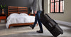 A man carrying the Tempur-Pedic Travel Set in its convenient rolling bag, walking past a modern bedroom with a wooden bed frame and white bedding, highlighting the portability and sleek design of the travel-friendly sleep solution.